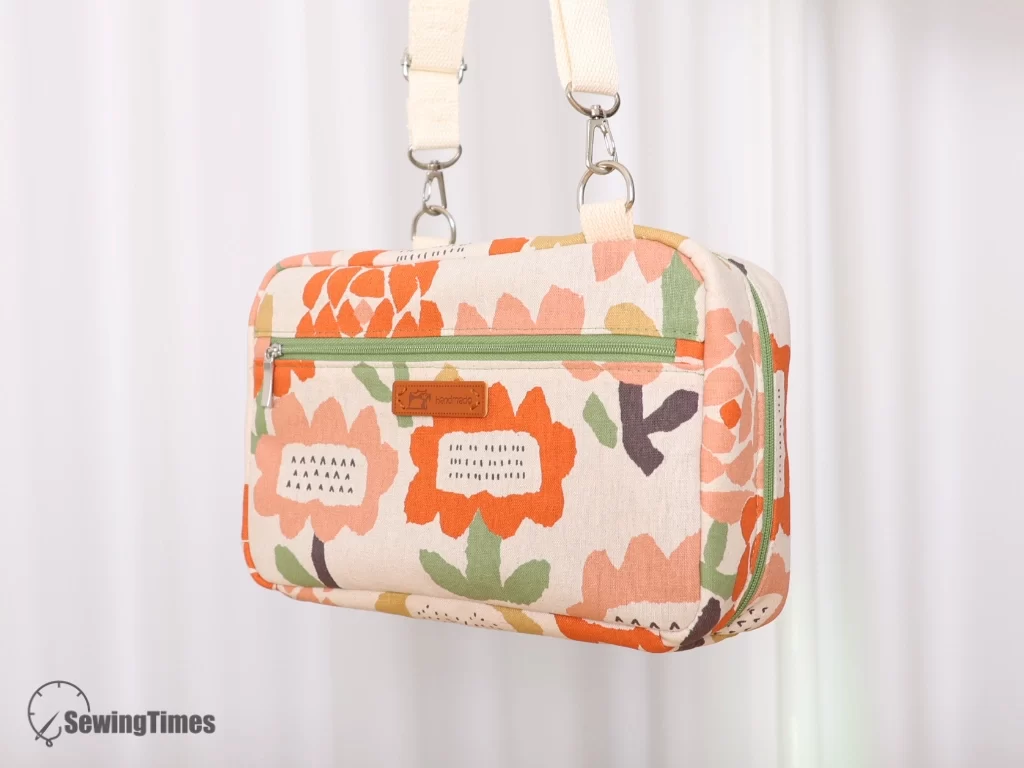 Front view of a handmade floral print accordion bag hanging by its off-white canvas webbing strap, featuring a green zipper pocket on the front and a small leather handmade label.