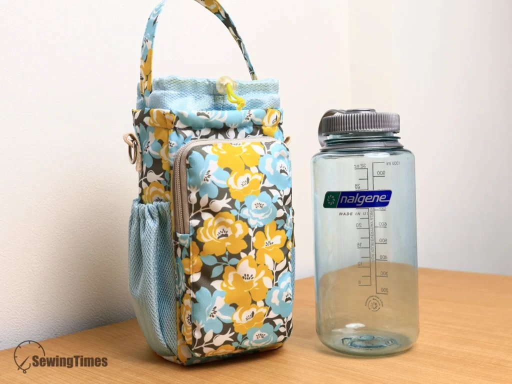 Handmade floral pattern water bottle carrier standing next to a clear 1-liter Nalgene water bottle on a wooden surface for size comparison.