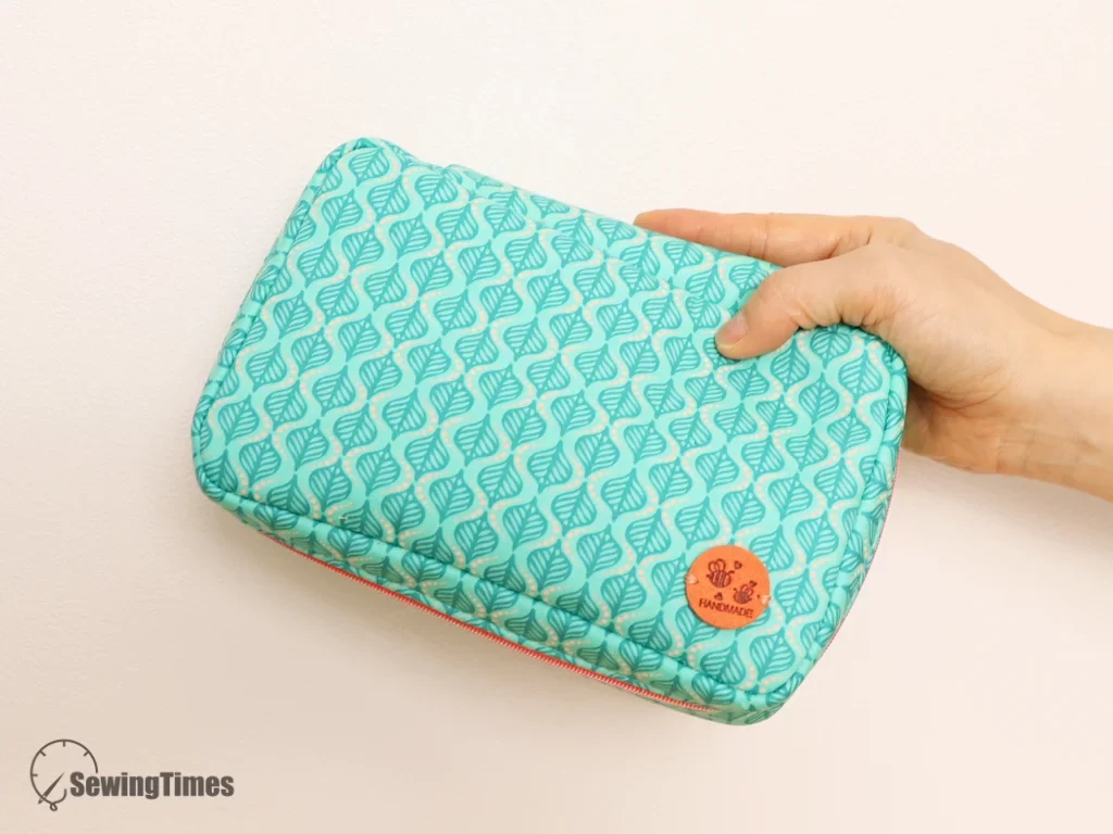 A hand holding a closed teal patterned rectangular makeup pouch, showcasing its compact size and pink zipper detail.