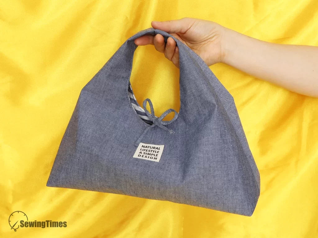 A person holding a handmade blue denim triangle bag with a minimalist white label, showcasing the unique geometric design and scale.