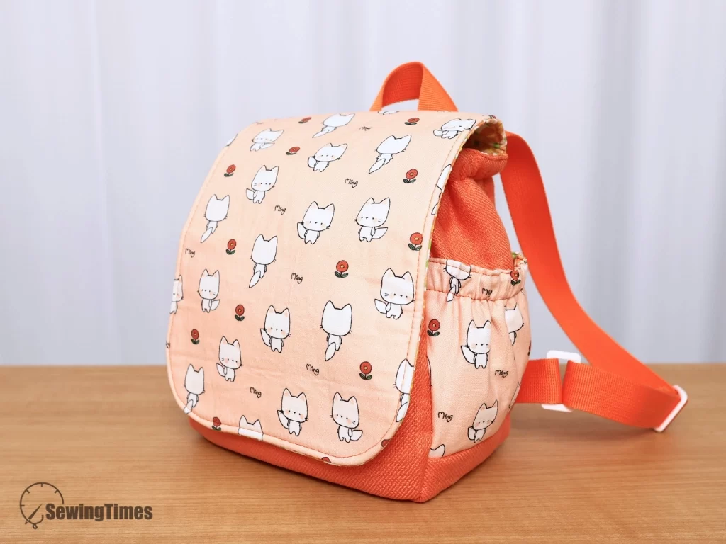 Front angle view of a small peach cat-print flap backpack for toddlers featuring adjustable straps and elastic side pockets.