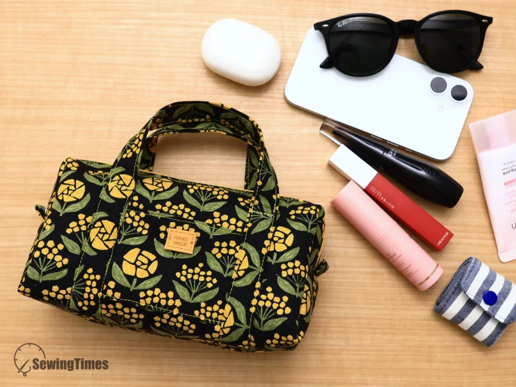 Black floral print mini duffle bag pouch surrounded by daily items like sunglasses, phone, and makeup.