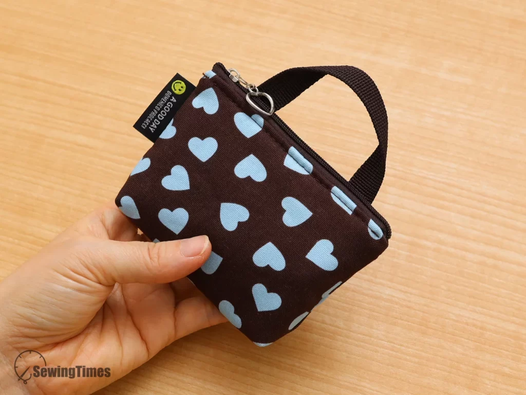 Close-up of a hand holding a mini zipper pouch with a light-blue heart pattern on a brown background, showcasing the main zipper pull with a silver ring and a fabric loop attachment.