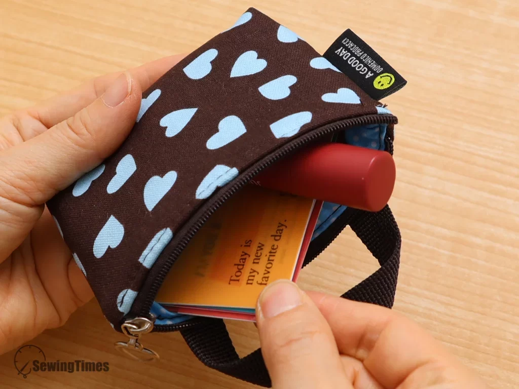 Hands are shown placing orange and teal credit cards and a red lip tint tube into the open brown mini zipper pouch, which is lined with a blue polka dot fabric.