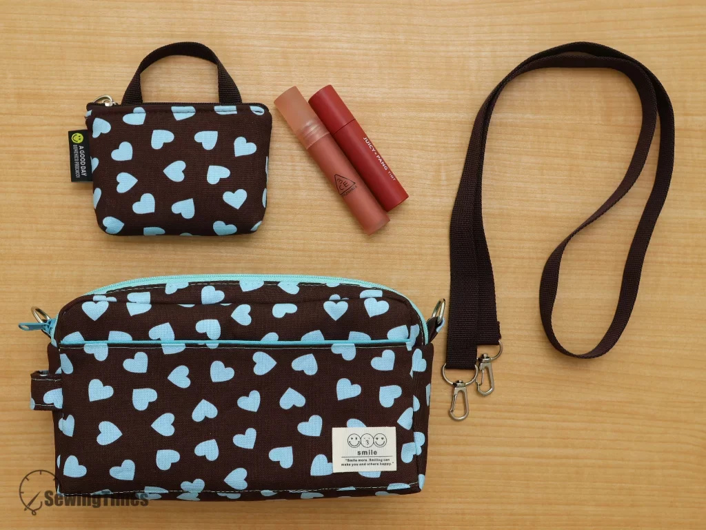A flat lay composition on a wood grain surface featuring a mini heart pattern zipper pouch (top left), two red lip tints, a matching larger heart pattern handbag (bottom center), and a detached brown fabric strap.