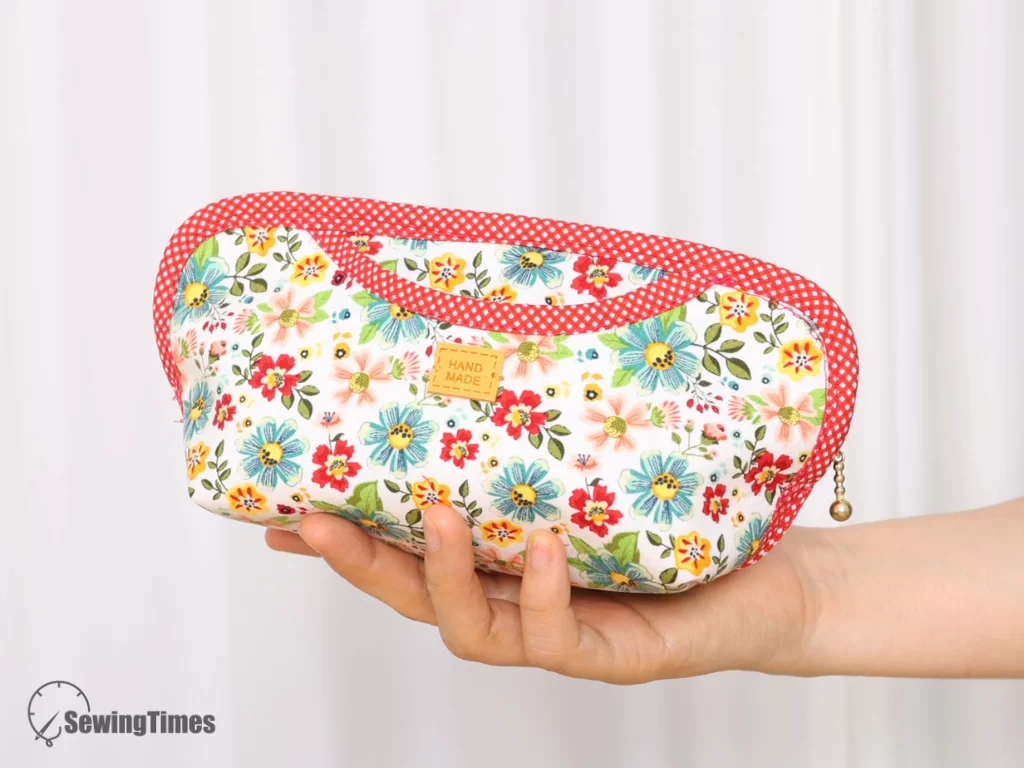 A handmade wide-open pencil case with a floral pattern fabric body, featuring a curved outer pocket with red and white polka dot bias binding, held in a person's hand.