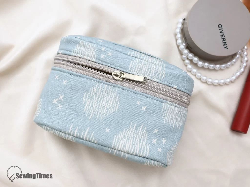 Front view of a closed light blue fabric oval zipper pouch next to a compact mirror and lipstick.