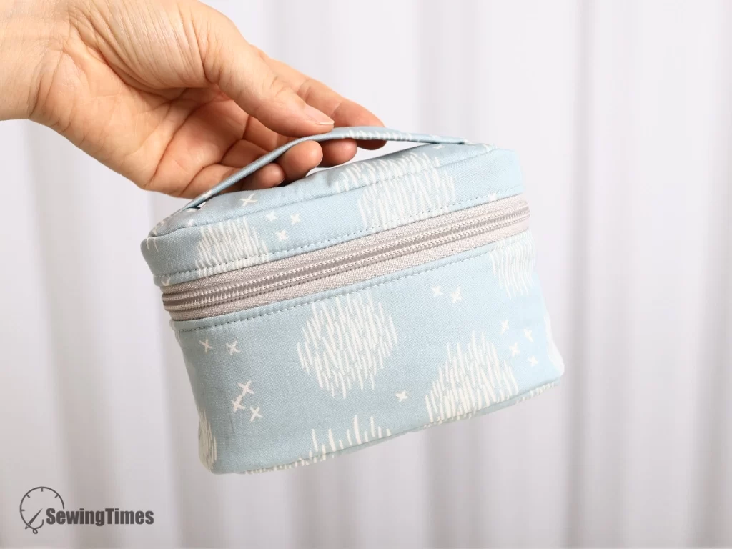 Hand holding a light blue handmade oval zipper pouch by its top handle against a neutral background.
