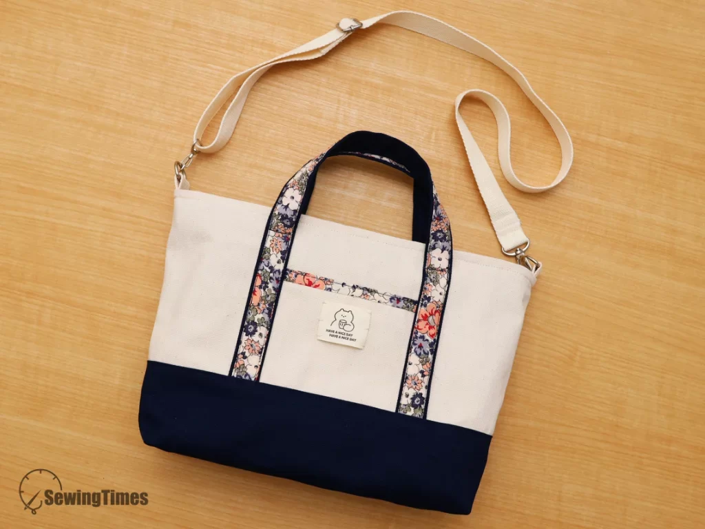 A cream canvas tote bag with an adjustable crossbody strap, floral handle detailing, and a navy blue bottom lying flat on a wooden surface.